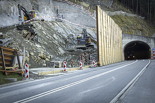 Mehrere Bagger spitzen in einer Baugrube Felsgestein ab; rechts im Bild befindet sich der Sisikoner Tunnel.