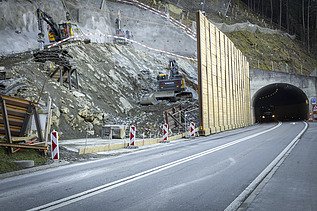Mehrere Bagger in einer Baugrube am Fels abspitzen, rechts ist der Sisikoner Tunnel zu sehen