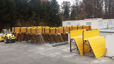  Ein Kran der STRABAG hebt an einem sonnigen Tag ein grosses Brückenelement. Im Hintergrund stehen mehrere Personen auf der Baustelle, neben einem Wohngebäude.