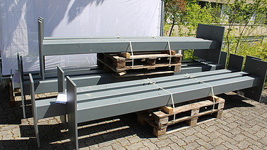 Mehrere graue Metallträger liegen auf Holzpaletten auf einem gepflasterten Boden, im Hintergrund ist ein Bauzaun