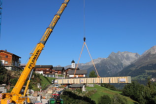 Ein Pneukran hebt einen langen Holzträger über ein Bergdorf im Vordergrund ein Kranführer und Monteure, im Hintergrund sind die Alpen zu sehen.
