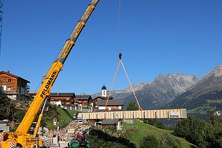 Ein Pneukran hebt einen langen Holzträger über ein Bergdorf im Vordergrund ein Kranführer und Monteure, im Hintergrund sind die Alpen zu sehen.