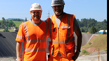 Zwei Männer in orangefarbener Bauausrüstung stehen lächelnd nebeneinander auf einer Baustelle mit Erdwall im Hintergrund.