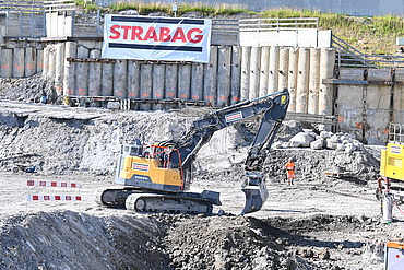  Ein gelber Bagger in einer Baugrube bei der Arbeit. Im Hintergrund sieht man ein STRABAG Banner 