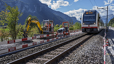 Ein Zug fährt auf einem Gleis durch eine Baustelle, rechts und links stehen Baumaschinen und Absperrungen. Im Hintergrund erheben sich hohe Berge unter blauem Himmel.