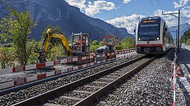 Ein Zug fährt auf einem Gleis durch eine Baustelle, rechts und links stehen Baumaschinen und Absperrungen. Im Hintergrund erheben sich hohe Berge unter blauem Himmel.