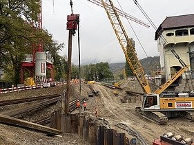 Baustelle am Flussufer: Sicht vom Wasser auf eine Staumauer, roten Kran und Spundwände der Baugrube.