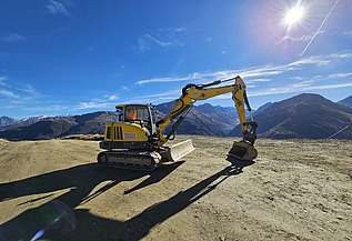 Gelber STRABAG Bagger auf einer geraden Sandfläche. Im Hintergrund blauer Himmel mit strahlendem Sonnenschein und einem Bergpanorama