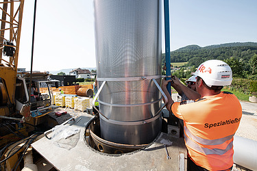 Ein Arbeiter steht neben einem grossen Metallrohr, das senkrecht in den Boden eingesetzt wird. Im Hintergrund sind Maschinen und Berge zu sehen.