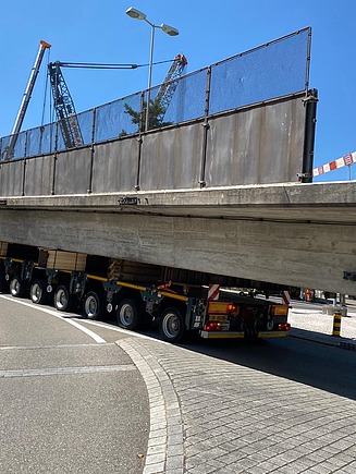 Ein massiver Beton-Brückenträger liegt auf einem langen Schwertransportfahrzeug, das über einen Kreisverkehr fährt. Im Hintergrund stehen Kräne.