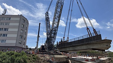  Ein Kran der STRABAG hebt an einem sonnigen Tag ein grosses Brückenelement. Im Hintergrund stehen mehrere Personen auf der Baustelle, neben einem Wohngebäude.