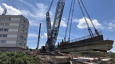  Ein Kran der STRABAG hebt an einem sonnigen Tag ein grosses Brückenelement. Im Hintergrund stehen mehrere Personen auf der Baustelle, neben einem Wohngebäude.