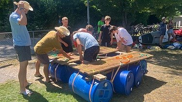 Lernende in Alltagskleider bauen ein Floss bei strahlendem Sonnenschein.