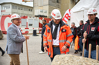 Mehrere Personen stehen bei einem Spatenstich zusammen. Eine Person im grauen Sakko spricht mit einem Arbeiter in orangefarbener Schutzkleidung und Helm, während andere Beteiligte mit Spaten im Hintergrund warten.
