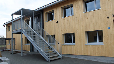 Asylunterkunft aus Holz, zweistöckig mit Metalltreppe und Geländer. 