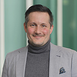 Kaufmännischer Bereichsleiter im Anzug lächelt in die Kamera. Im Hintergrund ist ein Lift zu sehen.