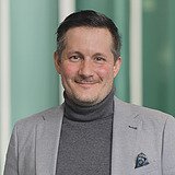 Kaufmännischer Gruppenleiter im Anzug lächelt in die Kamera. Im Hintergrund ist ein Lift zu sehen.