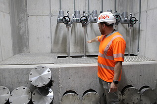 Ein Bauarbeiter in orangefarbener Sicherheitskleidung steht vor mehreren metallischen Rohranschlüssen in einer Betonkonstruktion und zeigt auf die installierten Komponenten.