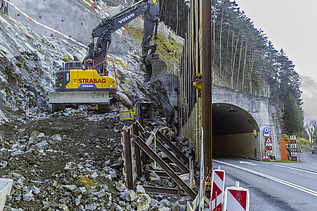 Ein gelber STRABAG Bagger arbeitet an einer Spundwand. Im Hintergrund sieht man einen Tunnel von der Axenstrasse und Bäume von einem Waldgebiet.