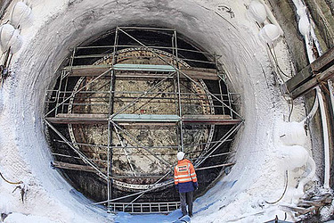 In der Mitte einer Bodenvereisung steht ein Bauarbeiter in Schutzkleidung vor einem Metallgerüst.
