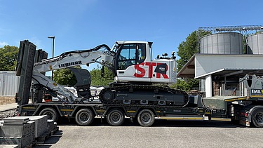 Anlieferung vom weisser STRABAG Liebherr-Raupenbagger, dieser steht verladen auf einem Tieflader-LKW