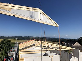 Rohbau der Labor-Erweiterung mit dem Einsatz eines Krans für eine weitere weisse vorgefertigte Wand und sichtbaren Holzbalken für das Dach. Im Hintergrund ländliche Umgebung mit Feldern, Bäumen und Hügeln.