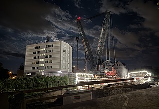 Nachtaufnahme einer STRABAG-Baustelle. Ein grosser Kran steht beleuchtet neben einem Wohnhaus unter dem klaren Nachthimmel.
