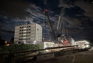 Nachtaufnahme einer STRABAG-Baustelle. Ein grosser Kran steht beleuchtet neben einem Wohnhaus unter dem klaren Nachthimmel.