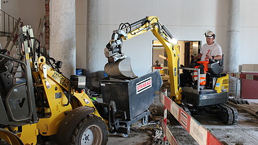 Bauarbeiter in STRABAG-Kleidung bedient einen kleinen Bagger in einem historischen Innenraum mit Säulen. Der Bagger leert Material in einen mit STRABAG-Logo versehenen Container. Links im Bild ist eine gelbe Baumaschine zu sehen. Die Baustelle ist mit Absperrbarrieren gesichert.