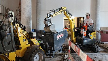 Bauarbeiter in STRABAG-Kleidung bedient einen kleinen Bagger in einem historischen Innenraum mit Säulen. Der Bagger leert Material in einen mit STRABAG-Logo versehenen Container. Links im Bild ist eine gelbe Baumaschine zu sehen. Die Baustelle ist mit Absperrbarrieren gesichert.