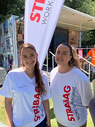 Zwei lächelnde Mitarbeitende in weissen STRABAG-T-Shirts stehen vor einem mobilen Stand.