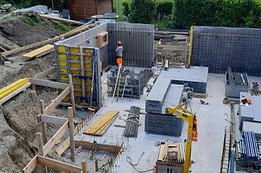 Eine Baugrube mit den Fundamenten und ersten Mauern eines 3-Familienhauses, ein Bauarbeiter bei der Arbeit ist ebenfalls zu sehen. 