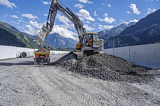 Ein Bagger verteilt Schotter auf einer Baustelle. Der Maschinenführer arbeitet auf einem grossen Steinhaufen, dahinter sind weisse Betonwände und die Berge zu sehen.