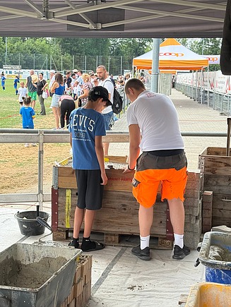 STRABAG Lernender in weissem STRABAG Shirt, erklärt einem Jungen wie man korrekt pflastert.