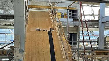  Ein Kran der STRABAG hebt an einem sonnigen Tag ein grosses Brückenelement. Im Hintergrund stehen mehrere Personen auf der Baustelle, neben einem Wohngebäude.