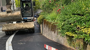 Bagger arbeitet am neu gebauten Trottoir; daneben ist eine rot-weiss markierte Absperrplatte auf der nassen Strasse zu sehen.