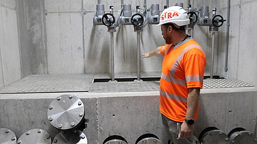 Ein Bauarbeiter in orangefarbener Sicherheitskleidung steht vor mehreren metallischen Rohranschlüssen in einer Betonkonstruktion und zeigt auf die installierten Komponenten.