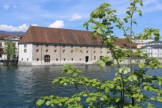 Historisches Landhaus Gebäude mit rotem Ziegeldach direkt an der Aare, mit vielen kleinen Fenstern und roten Fensterläden. Im Vordergrund grüne Zweige eines Baumes, im Hintergrund weitere Altstadtgebäude unter blauem Himmel.