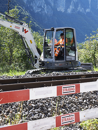 Ein kleiner weisser Bagger steht direkt neben den Bahngleisen und führt Arbeiten entlang der Strecke aus.
