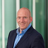 Raphael Wiederkehr Technischer Gruppenleiter  Technischer Gruppenleiter im Anzug lächelt in die Kamera. Im Hintergrund ist ein Lift zu sehen.