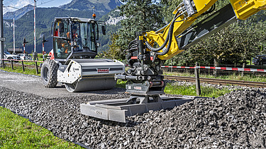 Ein gelber Bagger verdichtet frischen Schotter auf einer Strasse, während im Hintergrund eine weisse Walze bereitsteht. Rechts und links der Strecke sind Wiesen, Bäume und Schutzgeländer zu sehen. 
