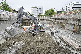 Ein Bagger steht auf Aushub in einer Baugrube hebt gerade die Schaufel an, im Hintergrund ist das Baustellenpersonal zu sehen