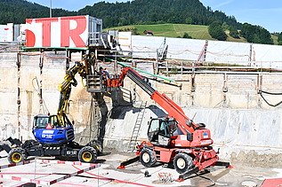 Zwei Baufahrzeuge in einer Baugrube bei der Arbeit. Im Hintergrund ein weisser STRABAG Container mit der Aufschrift STR in grossen roten Buchstaben.