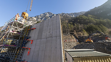 STRABAG-Baustelle im Gebirge: Betonarbeiten an einer hohen Stützmauer mit Kran und Bauführer:in auf dem Gerüst vor Bergkulisse.