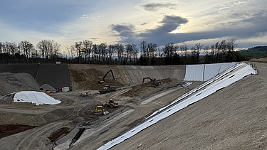 Blick über ein grosses Erdgefälle mit Baumaschinen unter einem wolkigen Himmel im Hintergrund sind Bäume zu sehen.