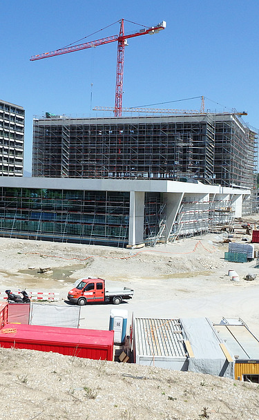 Grossbaustelle mit mehreren Gebäuden im Bau, darunter ein modernes, teilweise verglastes Gebäude im Vordergrund sowie ein grosses eingerüstetes Bauvolumen dahinter. Zwei rote Turmdrehkrane ragen in den Himmel. Auf dem Gelände befinden sich zahlreiche Baucontainer, Maschinen, Fahrzeuge und Baumaterialien. Rechts ein Baustellenweg, links Bauabsperrungen und Lagerflächen.