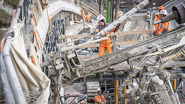 Nahaufnahme eines Bohrgeräts auf einer Baustelle. Mehrere STRABAG-Arbeiter in orangefarbener Schutzkleidung sind im Hintergrund an einer Betonwand tätig.