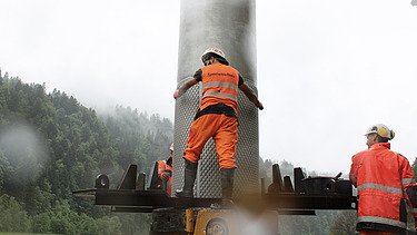 STRABAG Bauarbeiter in Warnkleidung steht auf einer Plattform und fixiert den Filterbrunnen. Im Hintergrund ist grauer Himmel und ein grüner Wald zu sehen.