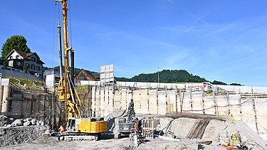 Ein grosser 15m hoher Bohrer in einer Baugrube bei der Arbeit. Im Hintergrund sieht man einen strahlend blauen Himmel. 