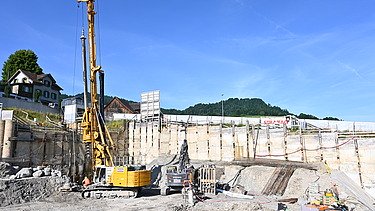 Ein grosser 15m hoher Bohrer in einer Baugrube bei der Arbeit. Im Hintergrund sieht man einen strahlend blauen Himmel. 